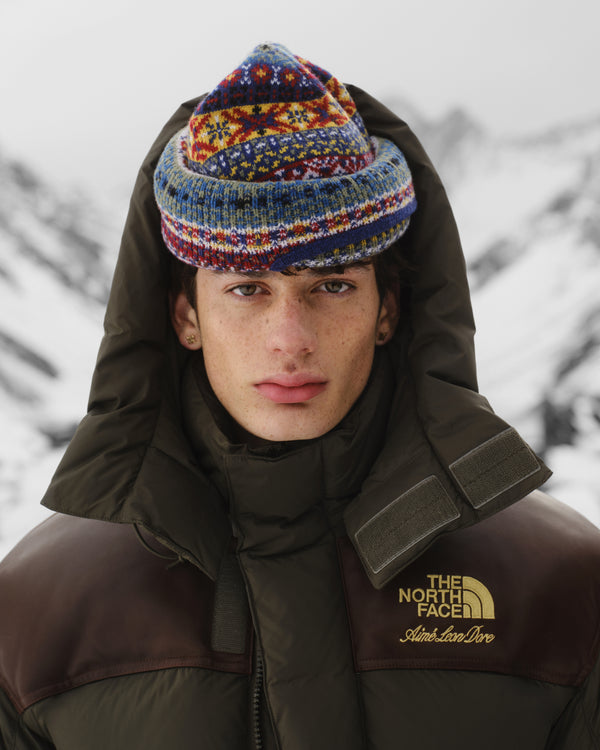 Young person in an ALD / The North Face Baltoro Jacket and knit hat stands in snowy mountains, facing the camera.