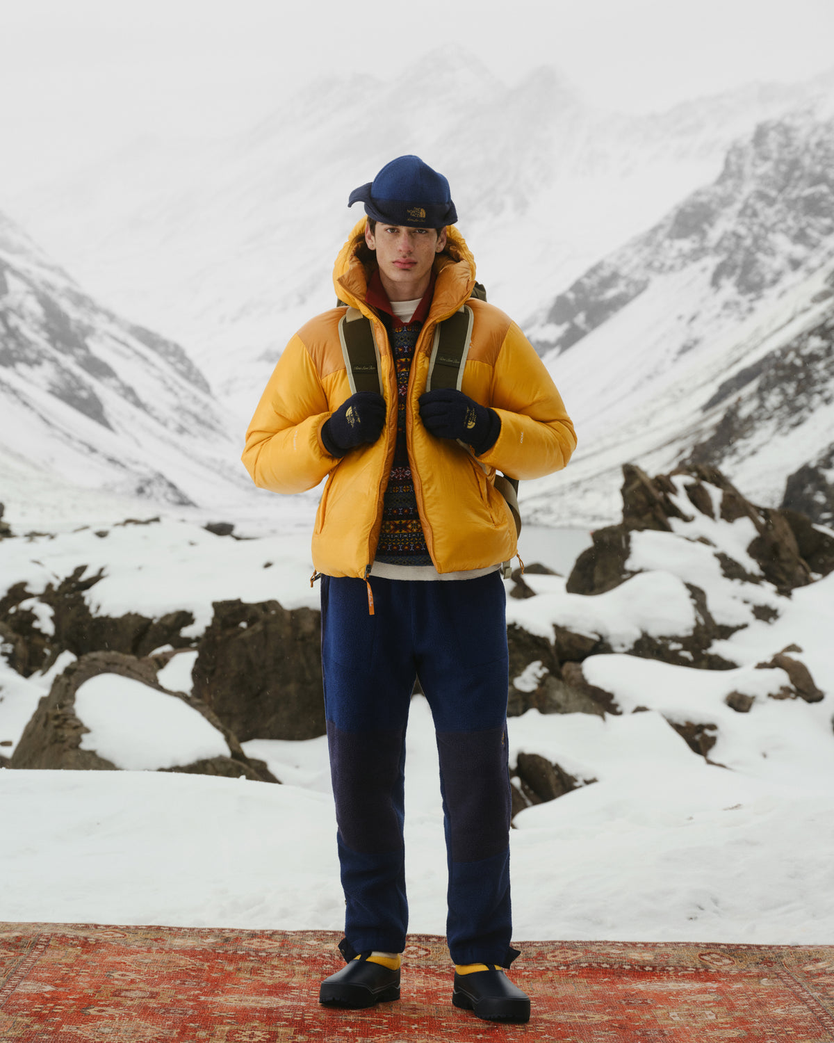 Someone in the ALD/The North Face Reversible Pertex® Nuptse Jacket stands on a rug in snowy mountains, wearing a backpack.