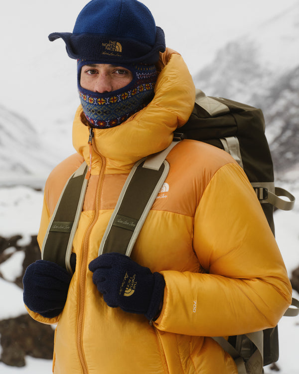A person wears the ALD / The North Face Fleece Earflap Hat, standing in snowy mountains with a backpack and face covering.