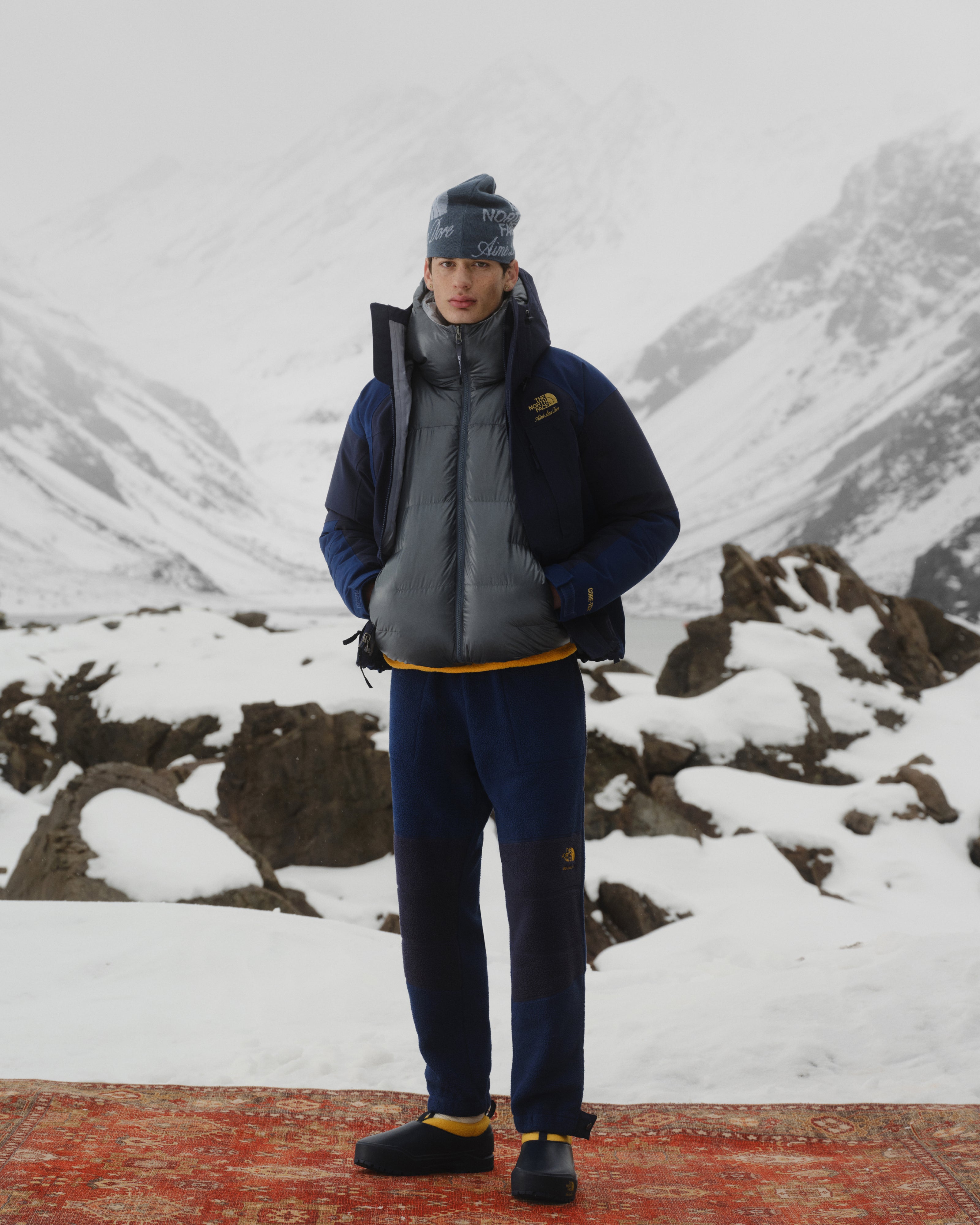 Person in ALD/The North Face GORE-TEX 2000 Mountain Jacket stands on a rug amid snowy peaks and rocks.