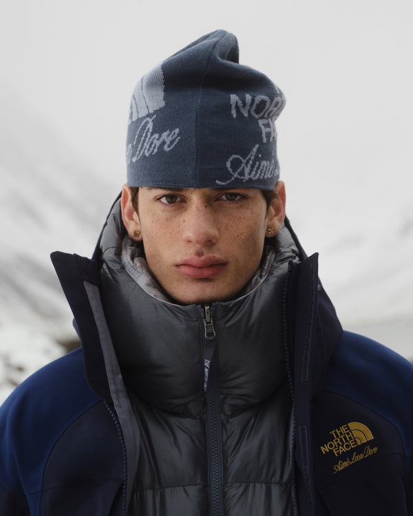 A man in a gray puffer and ALD / The North Face Jacquard Beanie stands outdoors with snowy mountains behind him.