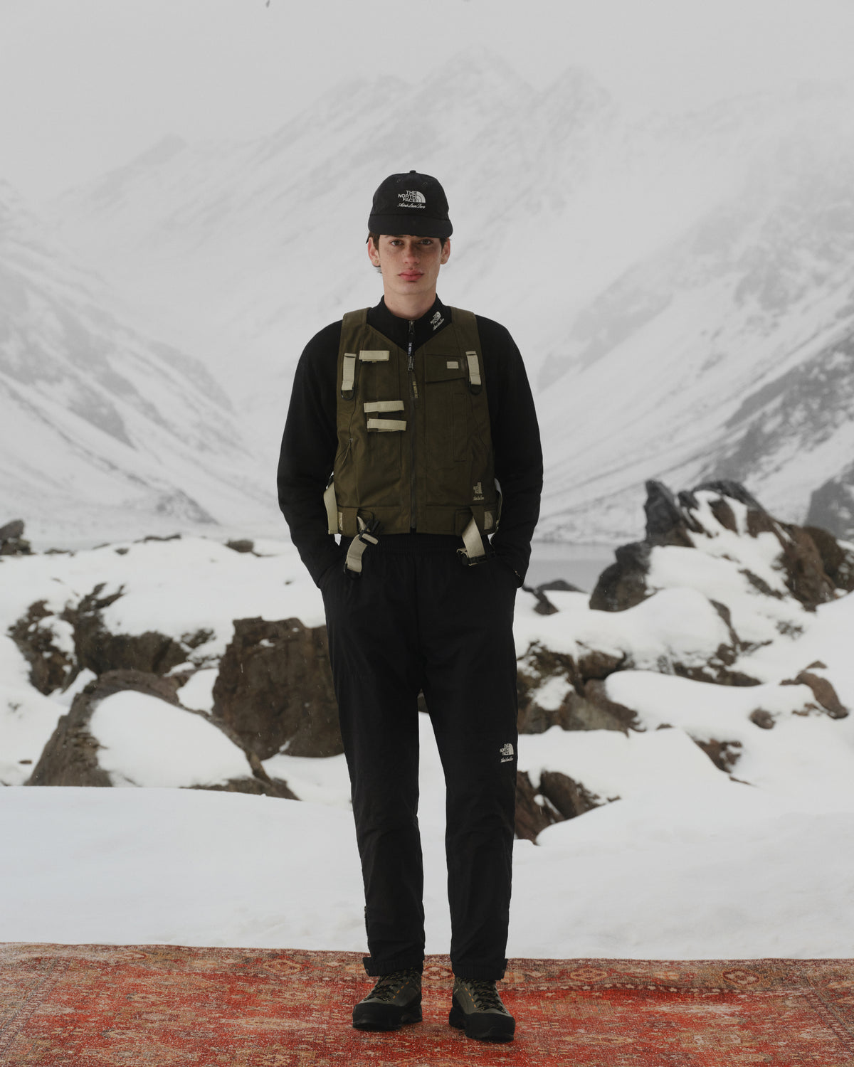 A person in outdoor gear wears the ALD / The North Face Nylon Logo Hat with snowy mountains and rocks behind.