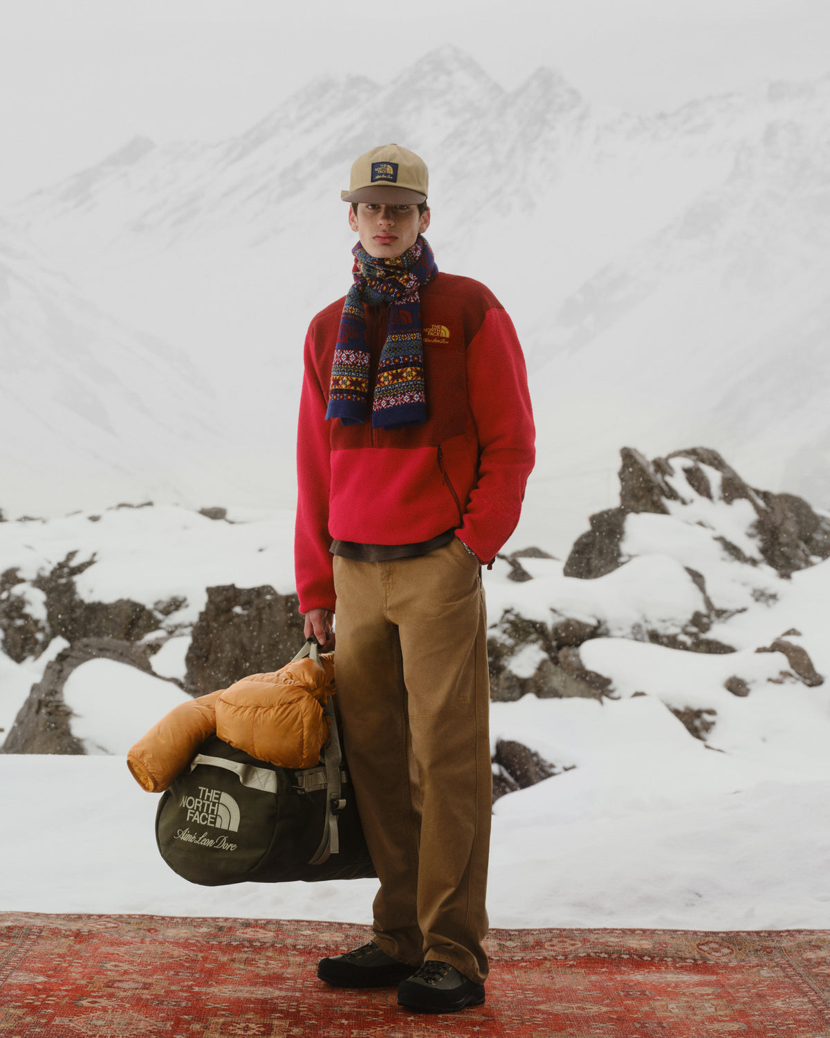 Man in red jacket and scarf stands on snowy mountain, holding ALD/The North Face Leather Brim Logo Hat, winter scene.