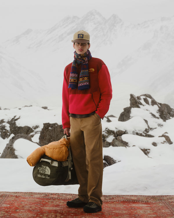A man in the ALD/The North Face ’95 Denali Half-Zip Fleece stands on a rug, snowy peaks behind, holding a duffel.
