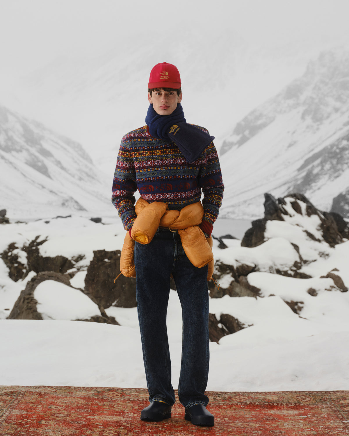 A man in winter clothes stands on a rug in snow, wearing the ALD/The North Face Denali Fleece Scarf.