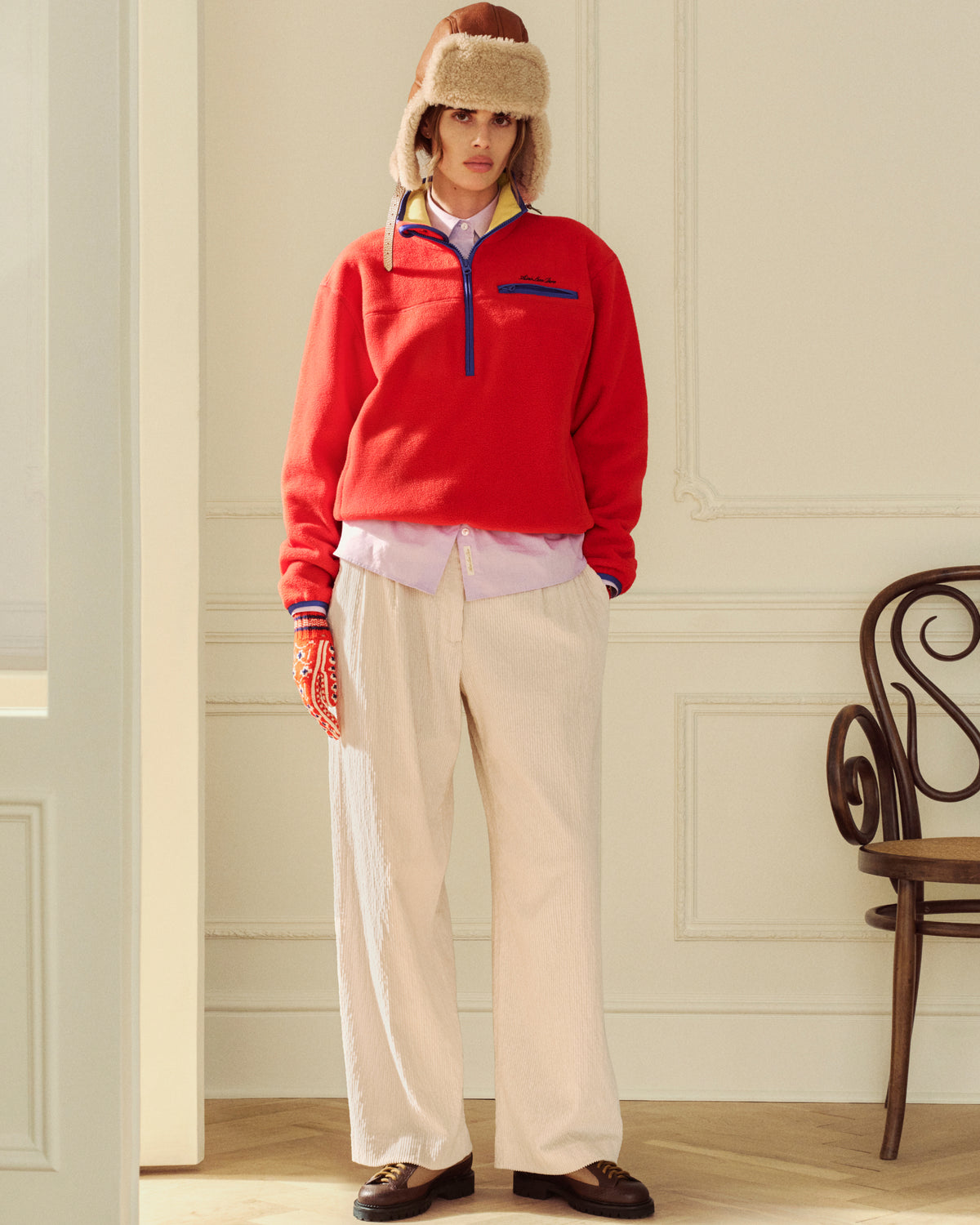 A person wears an aviator hat, red quarter-zip fleece, collared shirt, and cream Double Pleated Corduroy Trouser.