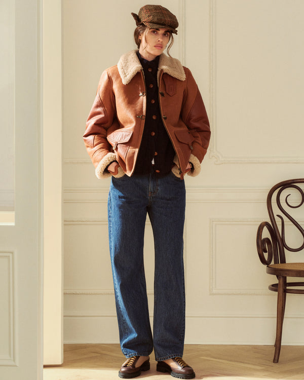 A person wearing a dark Twisted Rib Cardigan under a brown leather shearling jacket, blue jeans, and a flat cap.