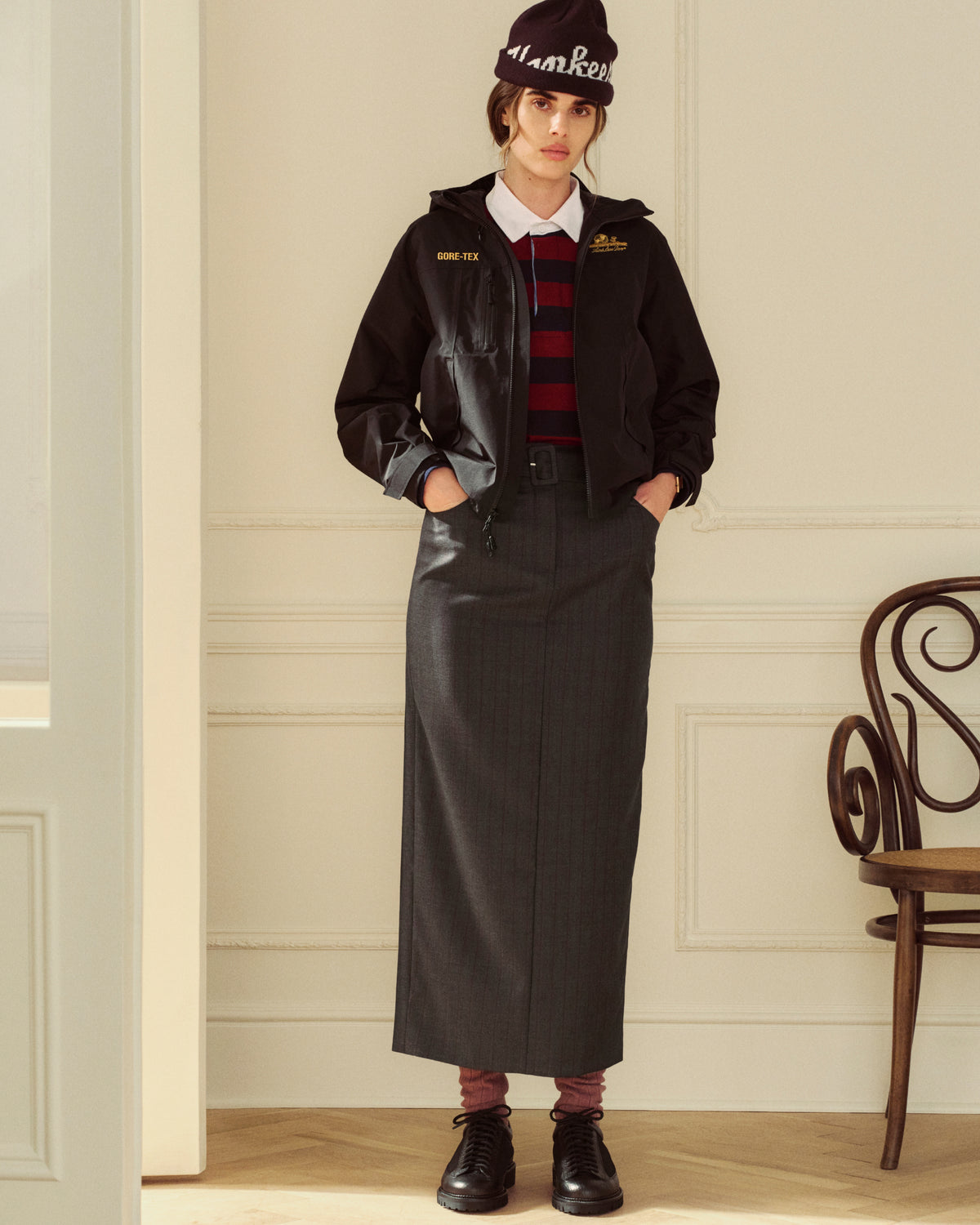 A person in a black jacket, Striped Script Rugby, and a dark skirt, paired with a knit beanie and boots.