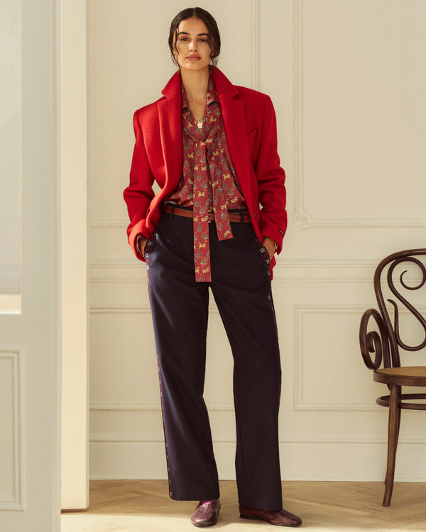 A person wearing a red blazer over an Archival Print Silk Tie Neck Blouse and dark trousers with side stripes.