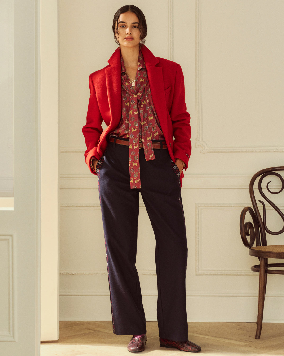 A person wearing a red Wool Sportcoat over a patterned pussy-bow blouse and navy trousers with side stripes.