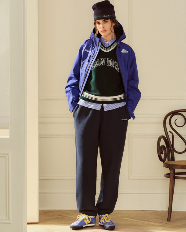 A person wearing a blue hooded jacket over a green v-neck sweater, Striped Button Down Poplin Shirt, and dark sweatpants.