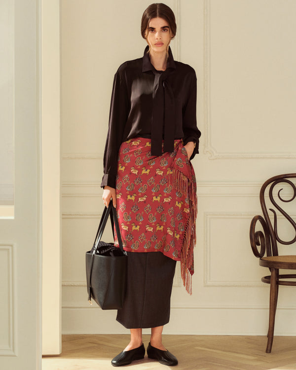 A woman in a black blouse and patterned red wrap skirt wears black Leather Ballet Flats while holding a black tote bag.
