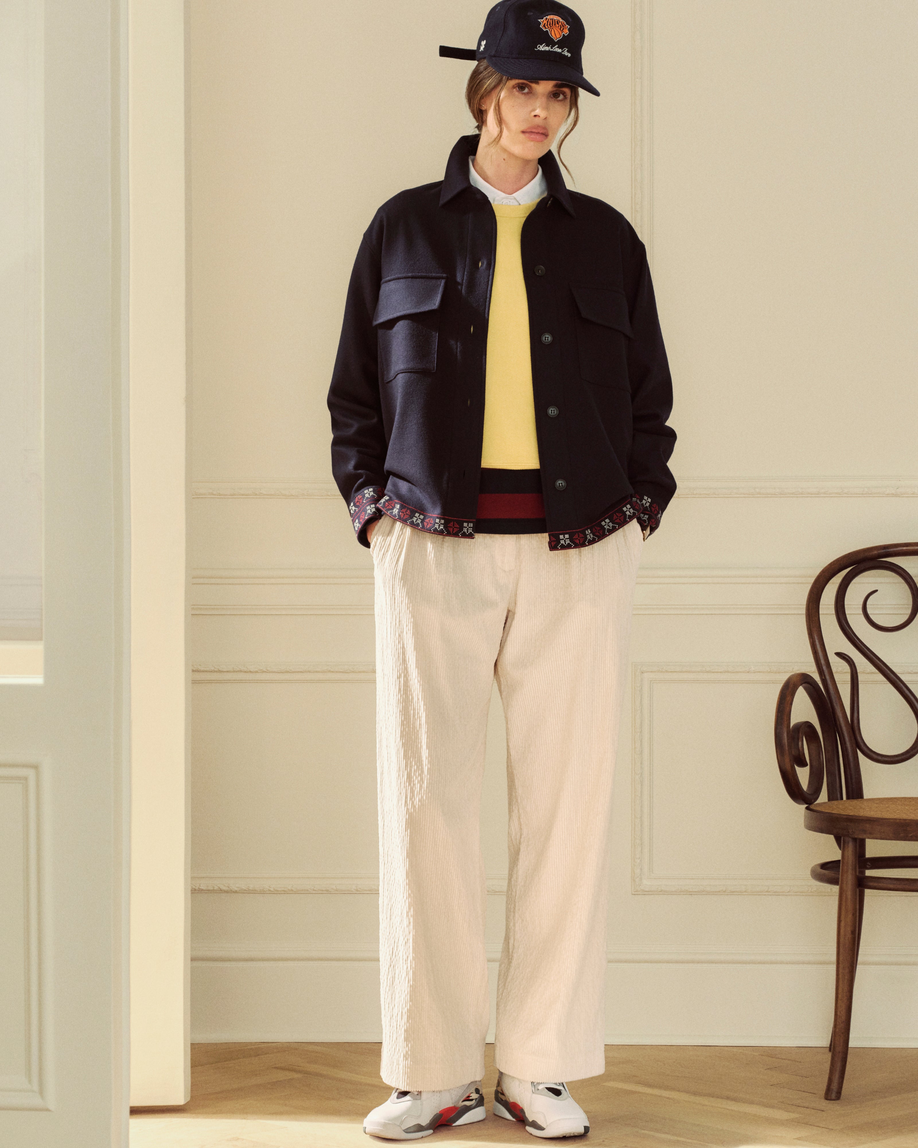 A person wears a yellow Crest Crewneck Sweatshirt under a dark jacket, light trousers, and a baseball cap.