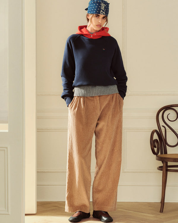 A model wears a navy sweatshirt over a red hoodie, tan wide-leg trousers, and ALD / New Balance MT2 sneakers.