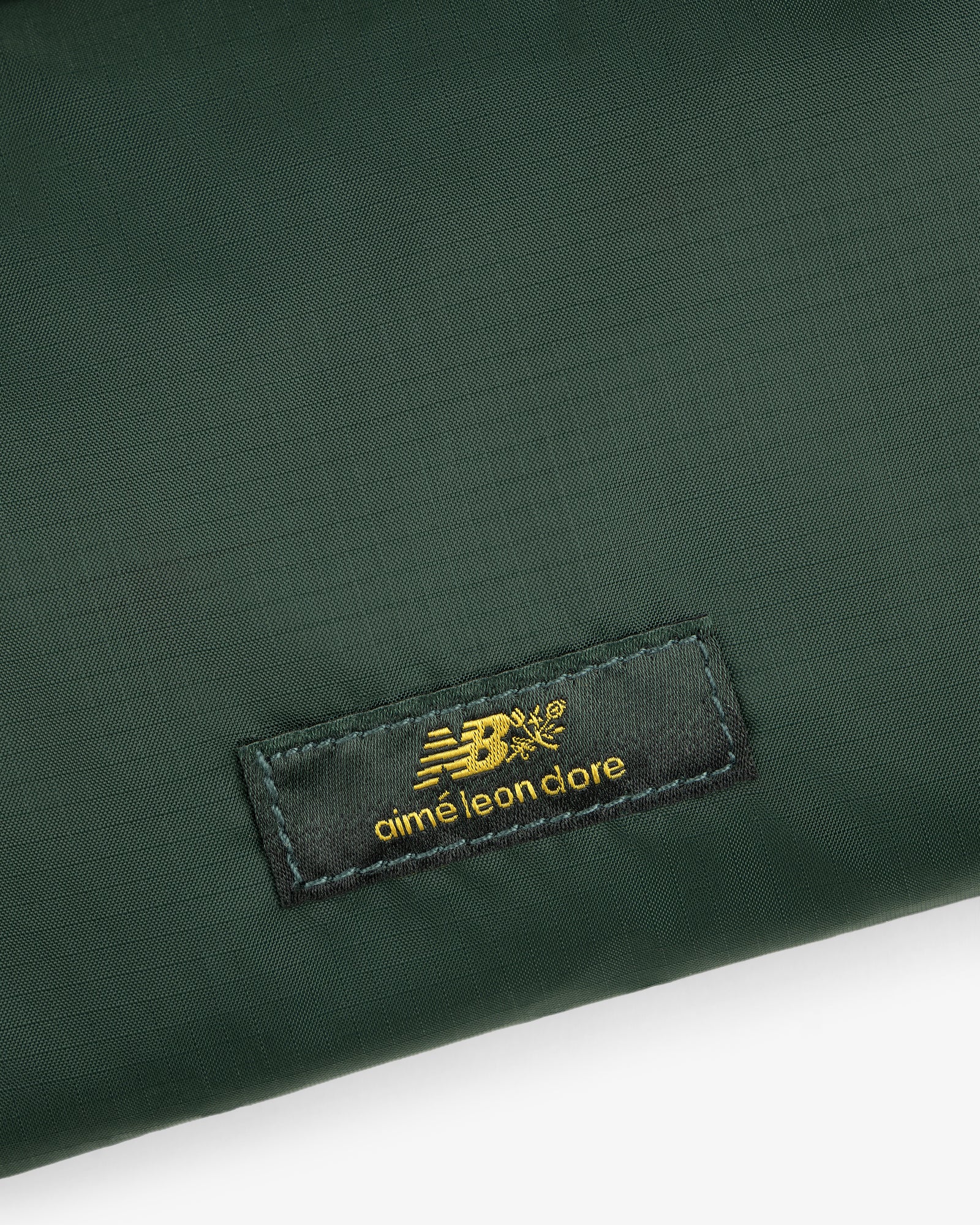 Close-up of a green ALD / New Balance Nylon Sling Pouch with a black patch featuring gold New Balance aimé leon dore text.