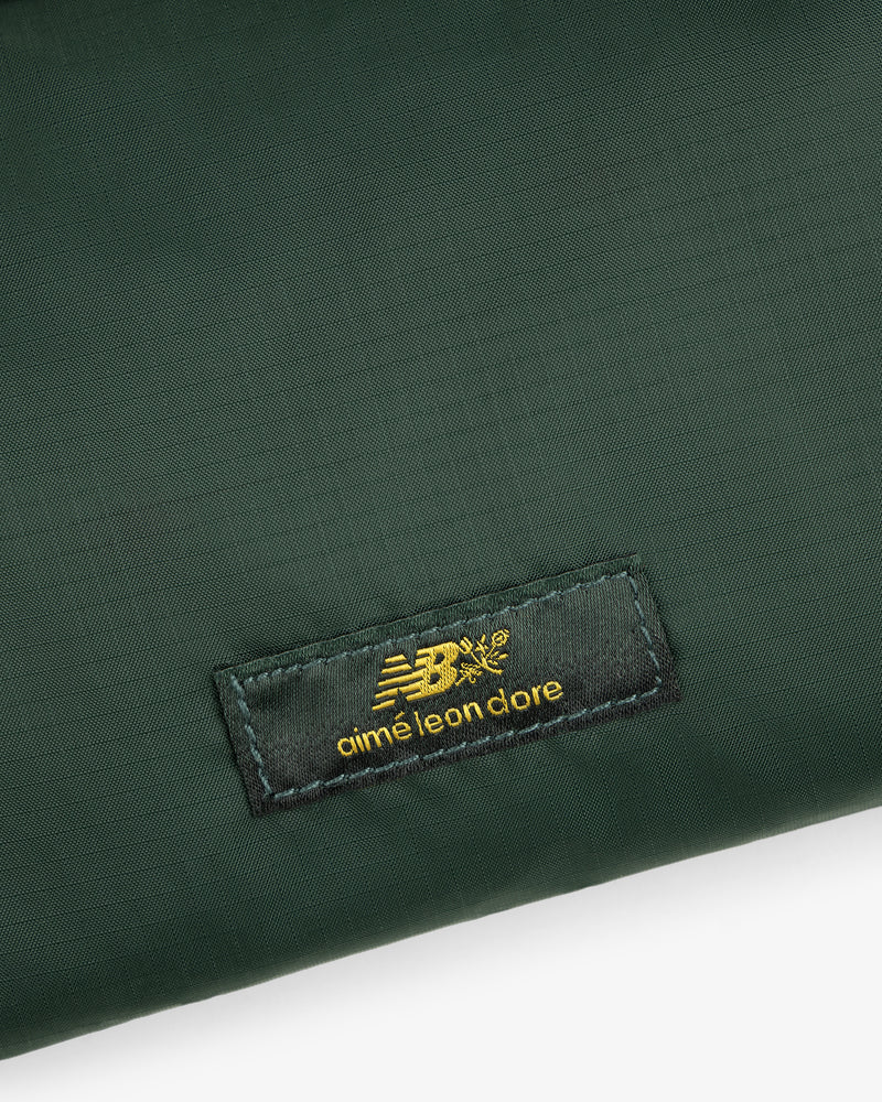 Close-up of a green ALD / New Balance Nylon Sling Pouch with a black patch featuring gold New Balance aimé leon dore text.