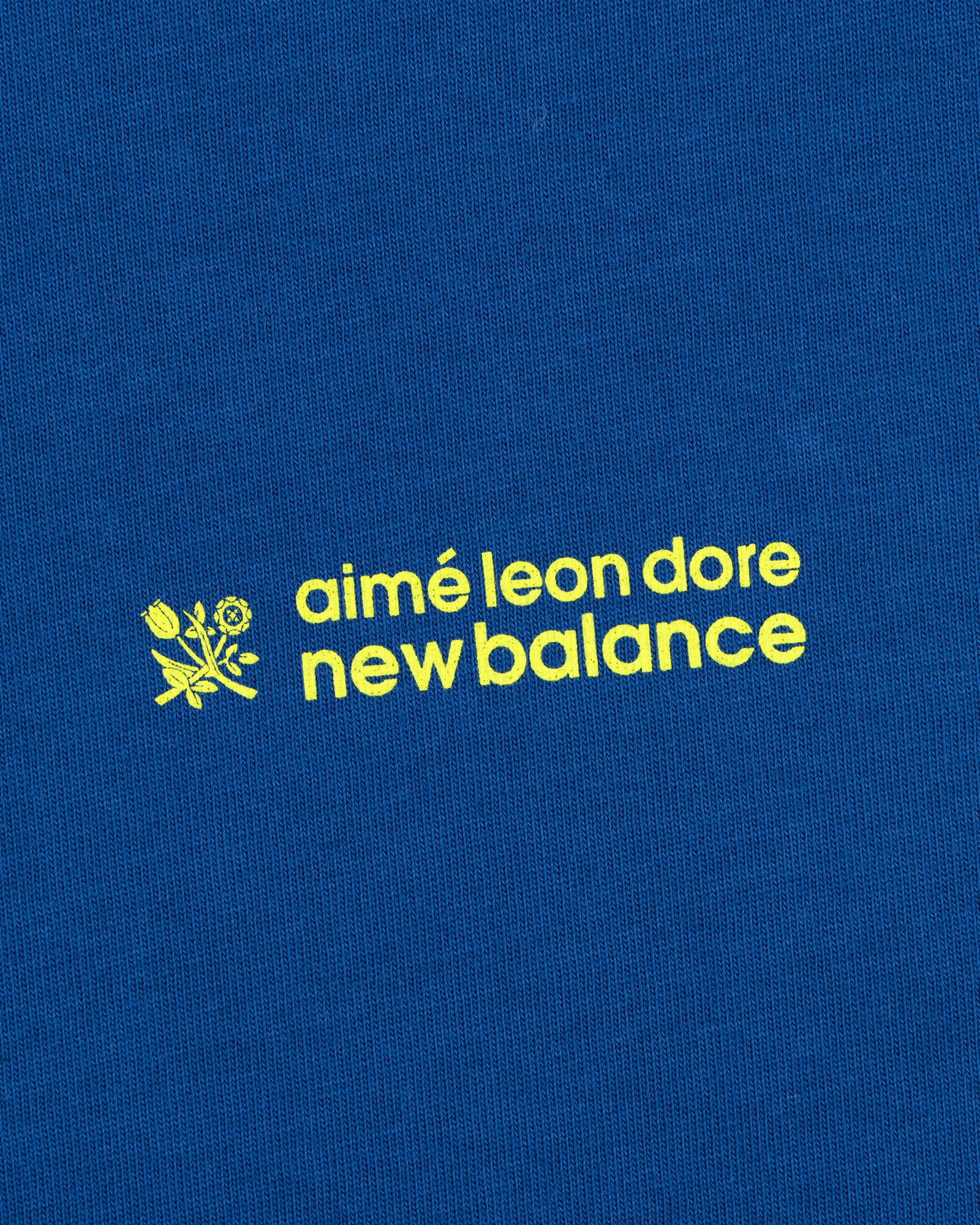 Yellow "ALD / New Balance Running Bib Tee" text and small flower graphic on a blue fabric background.