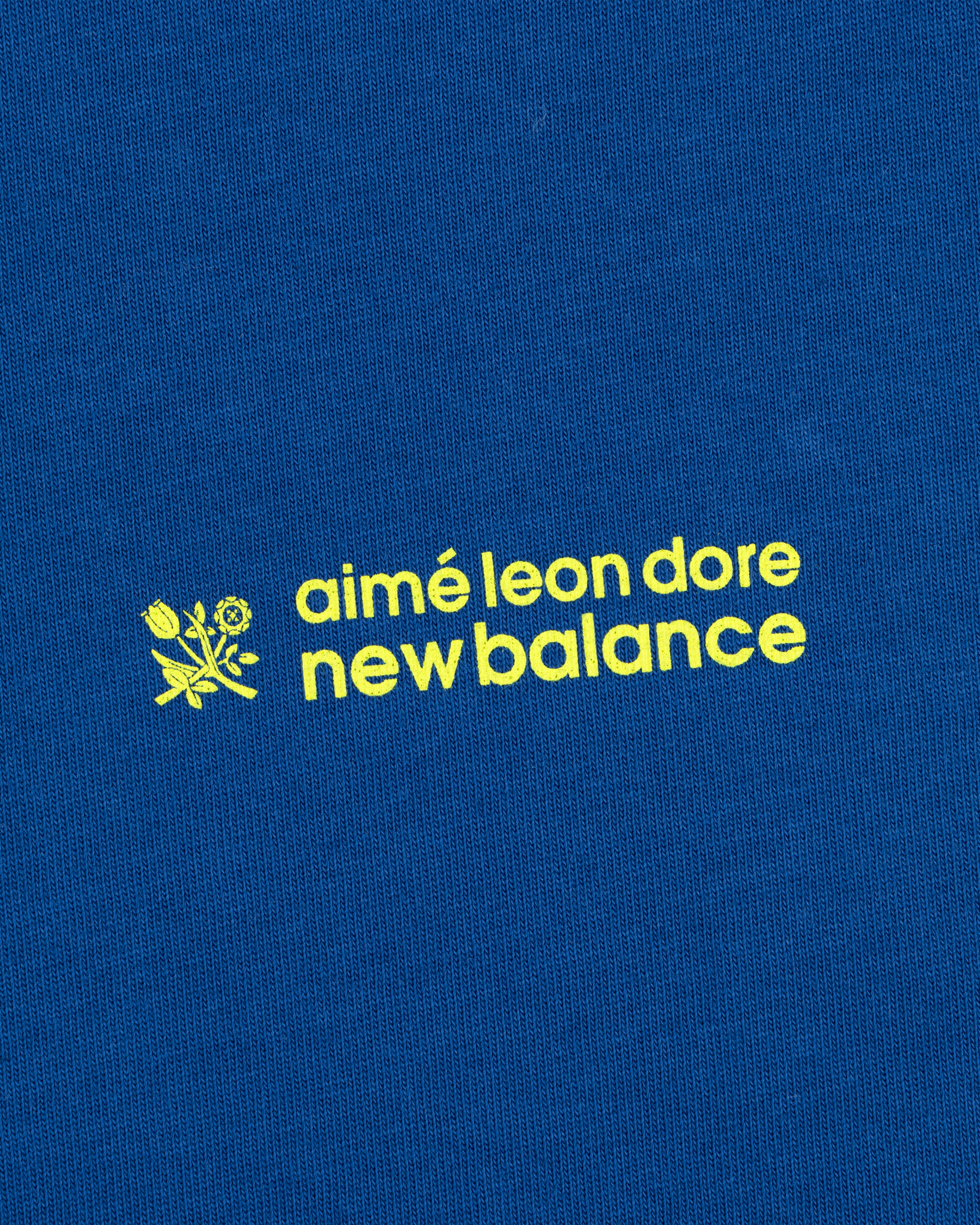 Yellow "ALD / New Balance Running Bib Tee" text and small flower graphic on a blue fabric background.