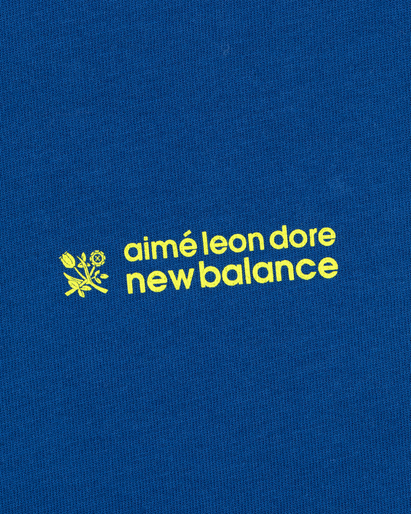 Yellow "ALD / New Balance Running Bib Tee" text and small flower graphic on a blue fabric background.