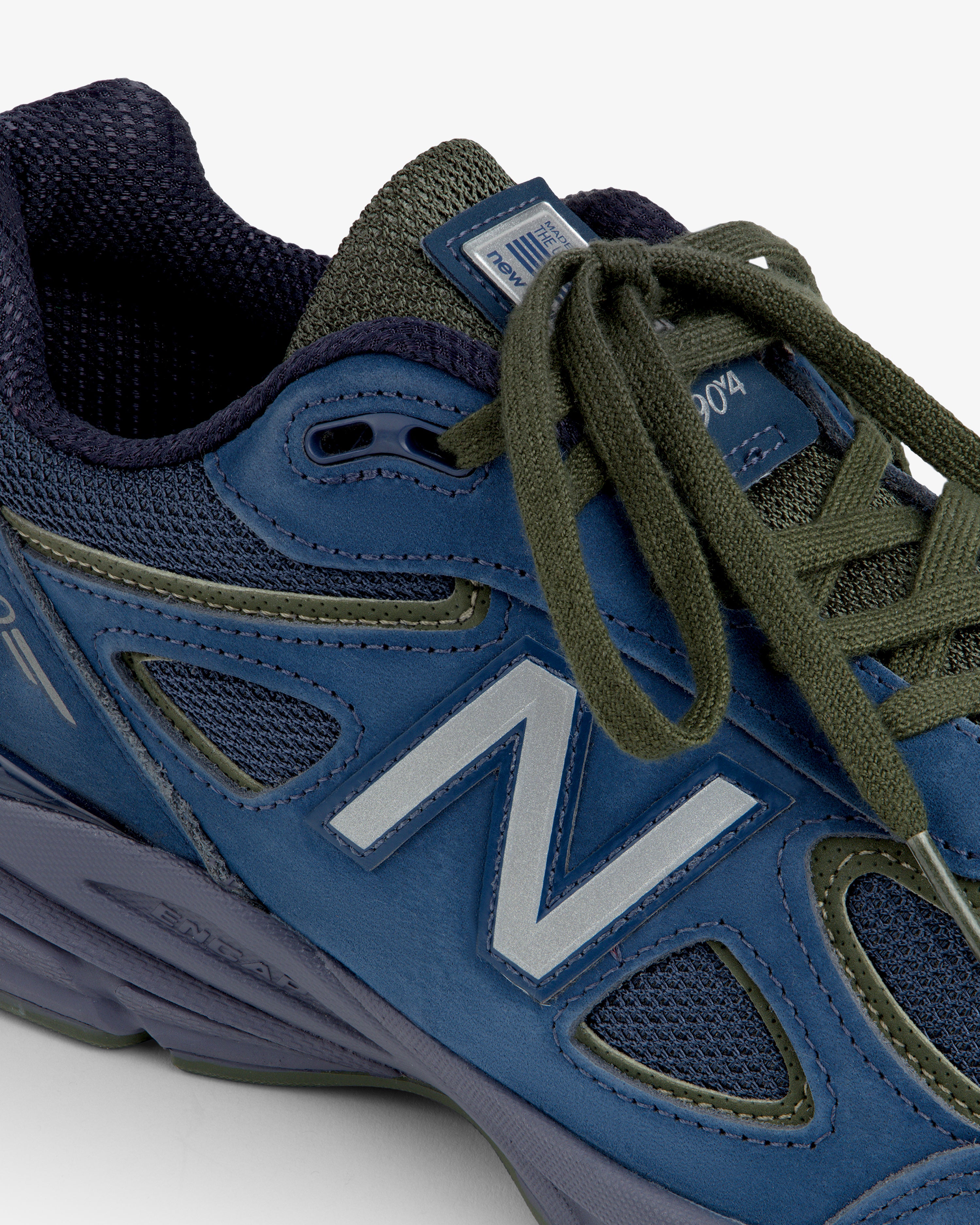 Close-up of a blue New Balance Made in USA 990v4 sneaker with olive green laces on a white background.
