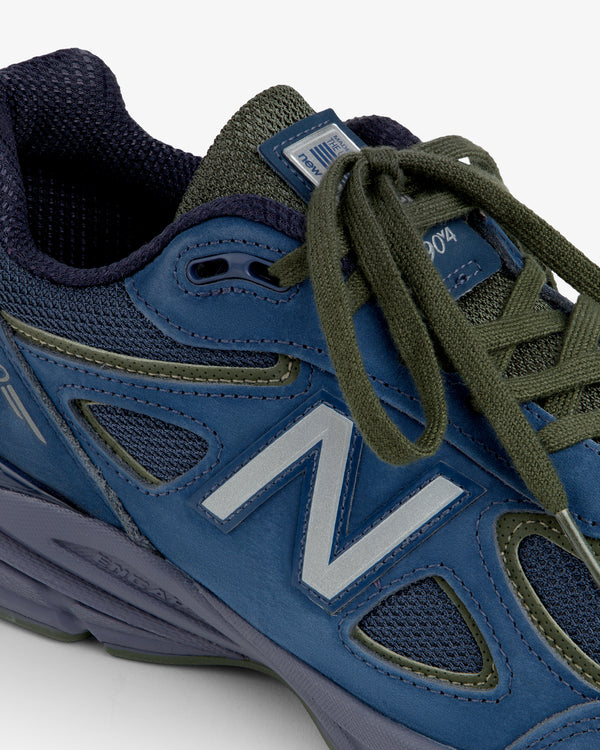 Close-up of a blue New Balance Made in USA 990v4 sneaker with olive green laces on a white background.