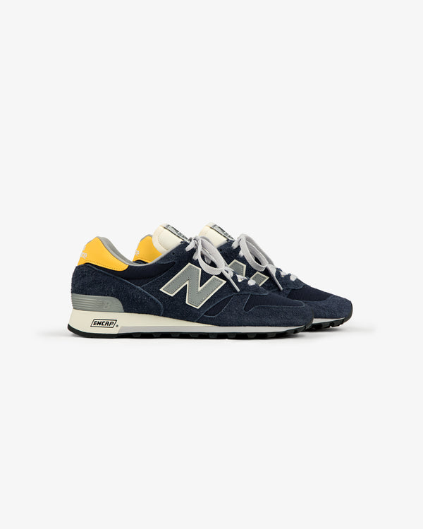 A pair of ALD / New Balance Made in USA 1300 navy blue and yellow sneakers with white laces on a white background.