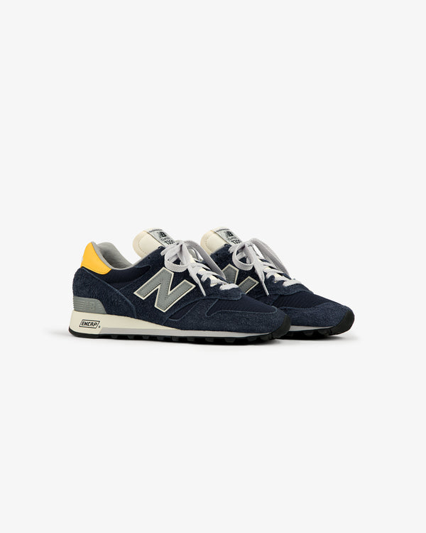 ALD / New Balance Made in USA 1300 sneakers in navy with white laces and yellow heel accents on a white background.