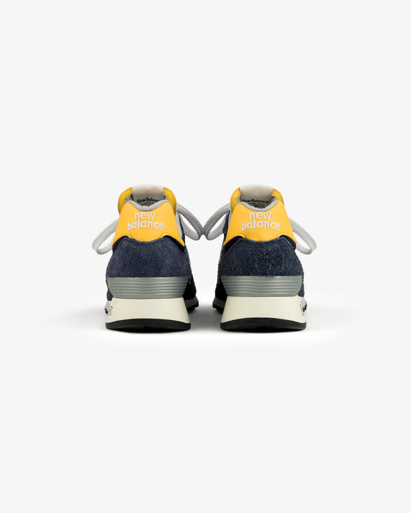 The ALD / New Balance Made in USA 1300 sneakers in navy and yellow, viewed from the back on a white background.