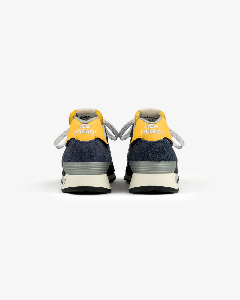 The ALD / New Balance Made in USA 1300 sneakers in navy and yellow, viewed from the back on a white background.