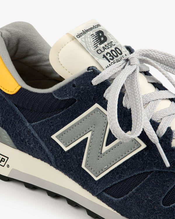 Close-up of ALD / New Balance Made in USA 1300 navy sneaker with gray laces, yellow heel, and N logo.