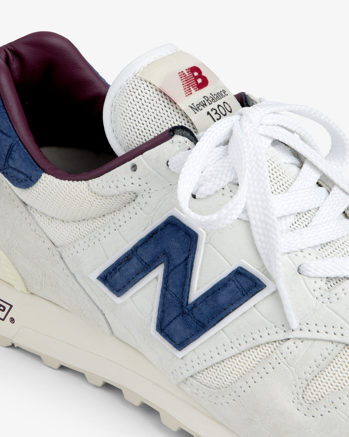 Close-up of a white New Balance Made in USA 1300 sneaker with blue logo, white laces, and maroon lining.
