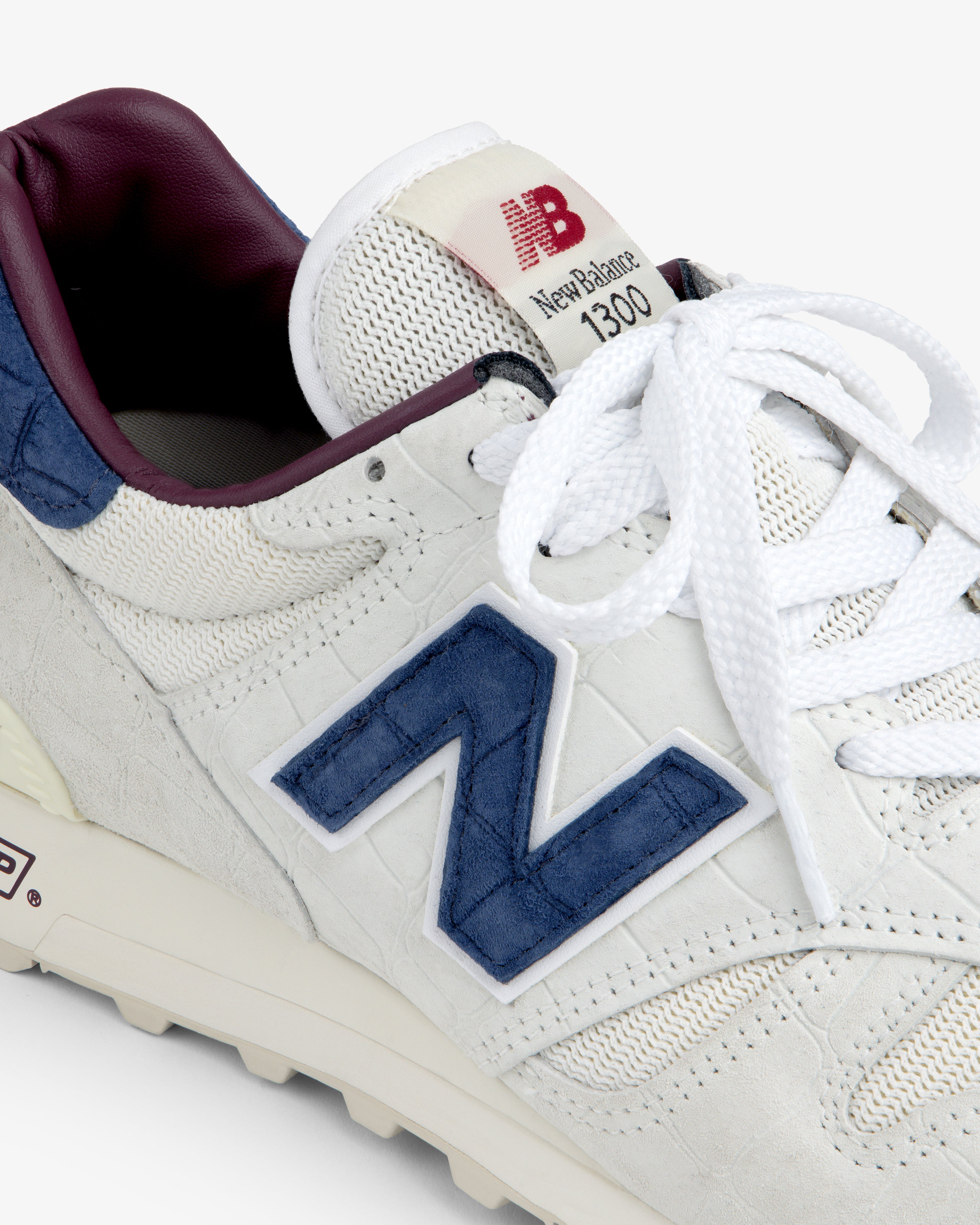 Close-up of a white New Balance Made in USA 1300 sneaker with blue logo, white laces, and maroon lining.