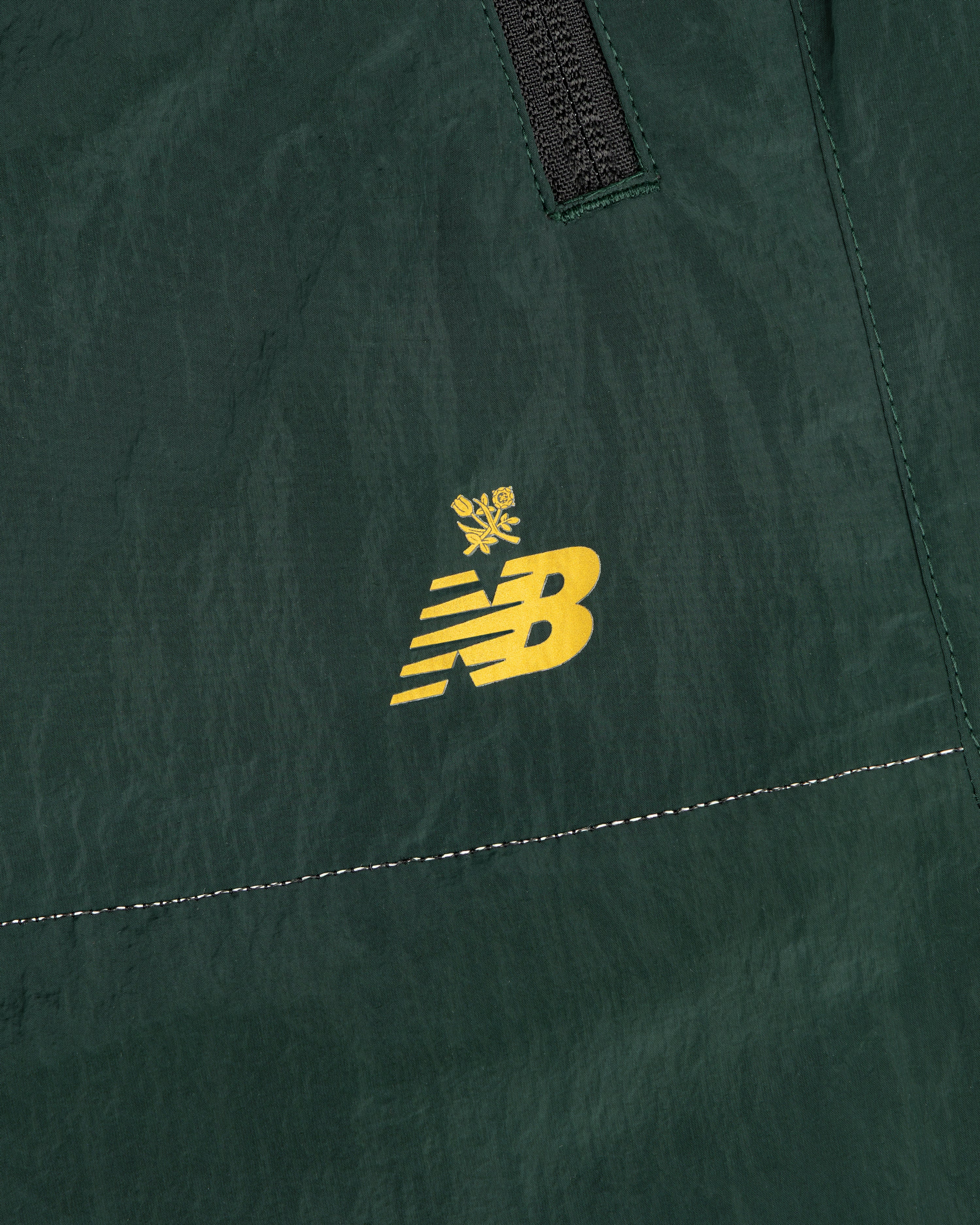 Close-up of ALD / New Balance Lightweight Nylon Warmup Pant in green with yellow NB logo, crown, seam, and zipper.