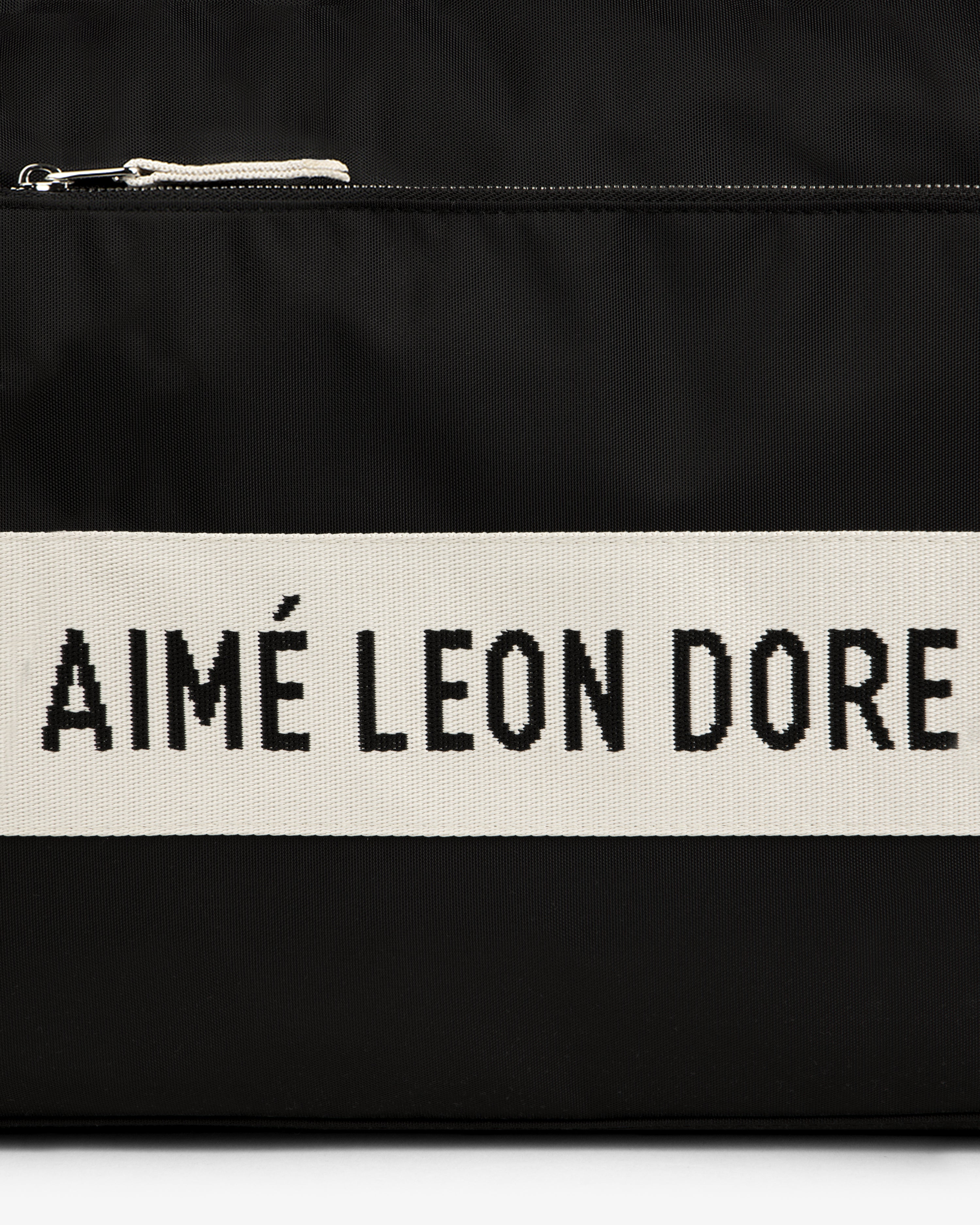 Black nylon duffle bag with a cream stripe featuring bold black AIMÉ LEON DORE text.