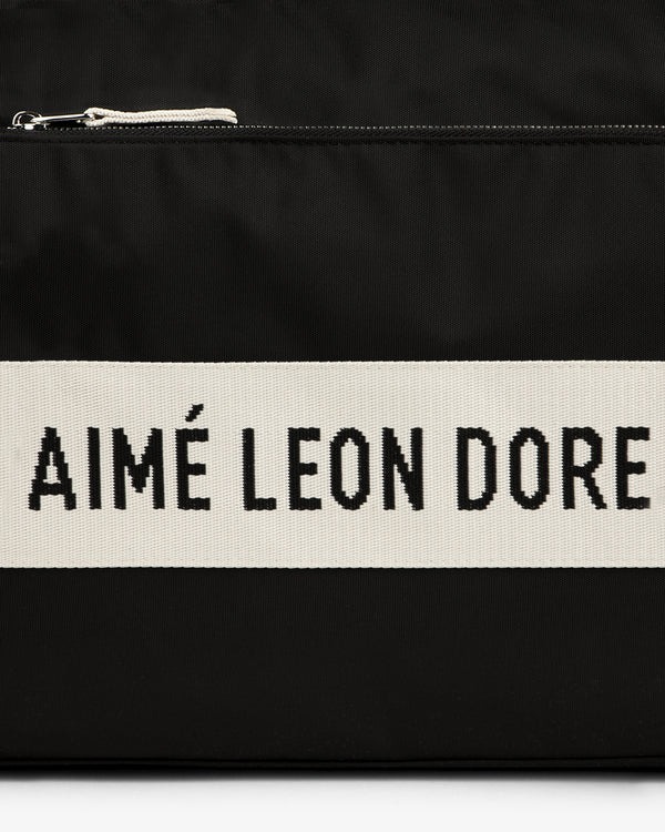 Black nylon duffle bag with a cream stripe featuring bold black AIMÉ LEON DORE text.