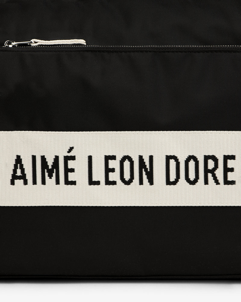 Black nylon duffle bag with a cream stripe featuring bold black AIMÉ LEON DORE text.