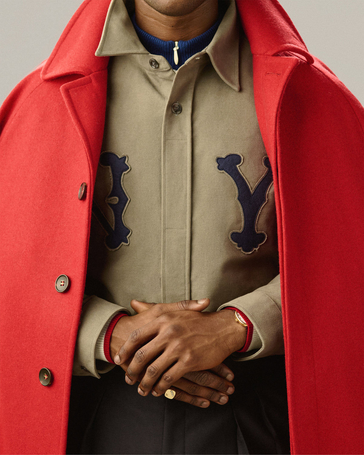 A person in a red coat over a tan shirt, hands clasped, wearing a 14Kt Gold Diamond Signet Ring and a watch.