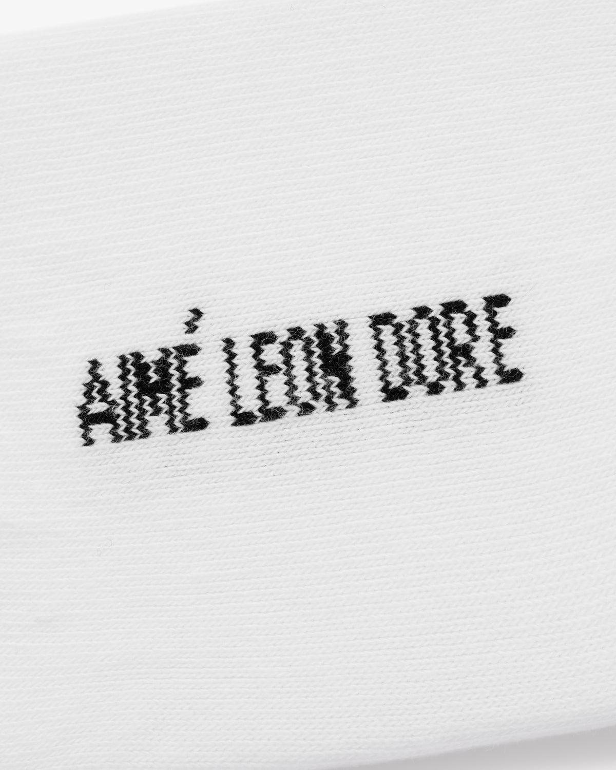 Close-up of white No-Show Sock 3-Pack with AMÉ LEON DORE in black, pixelated, embroidered text.