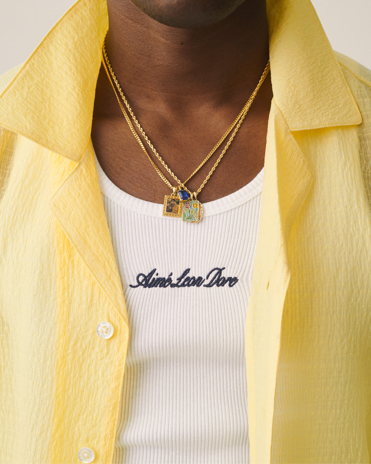 A person wears a yellow shirt over a white tank top with Aimé Leon Dore and two 14Kt Gold Mytilene Pendants.