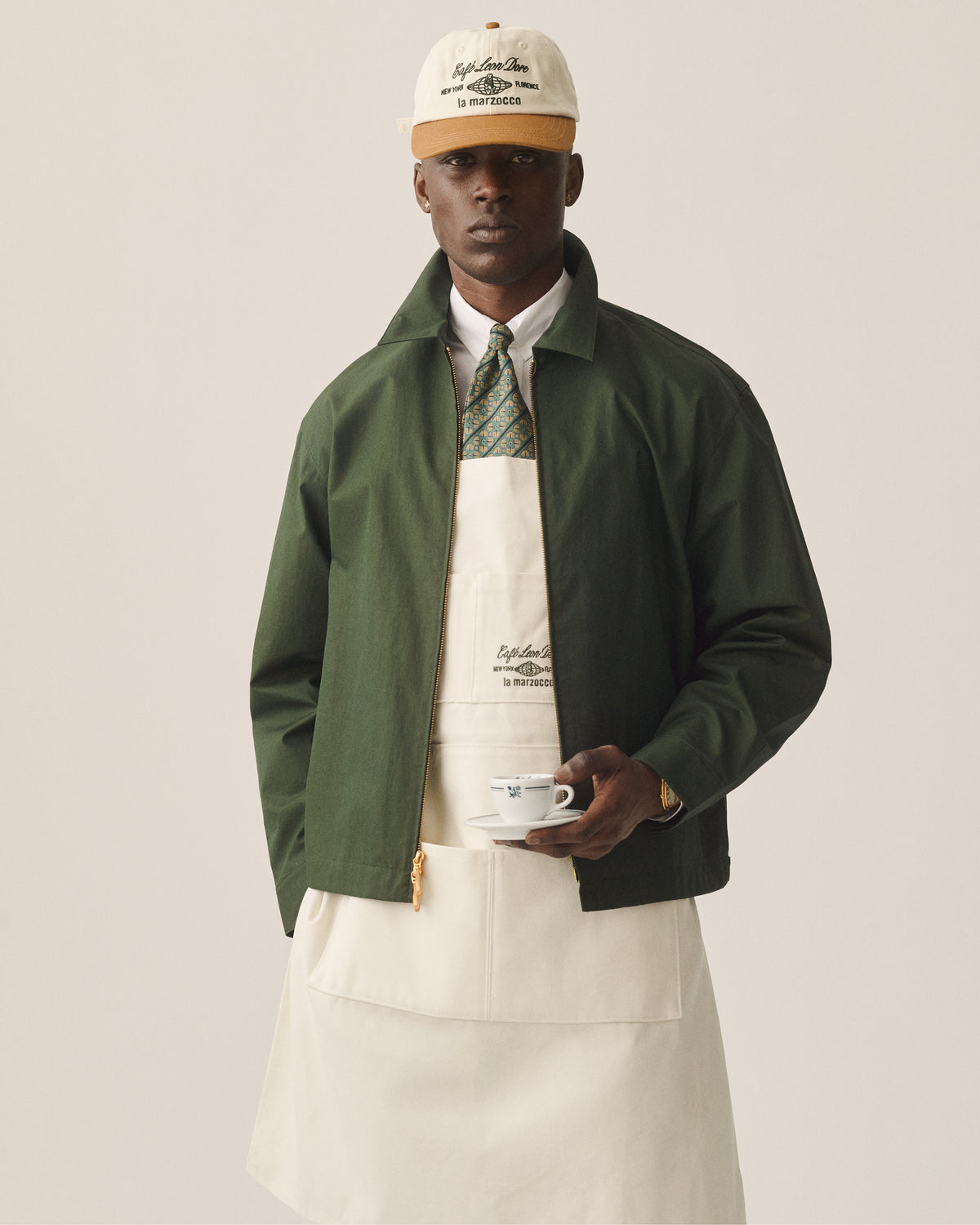 A man in a green jacket, tie, white apron, and Café Leon Dore / La Marzocco Hat holds a white cup and saucer.