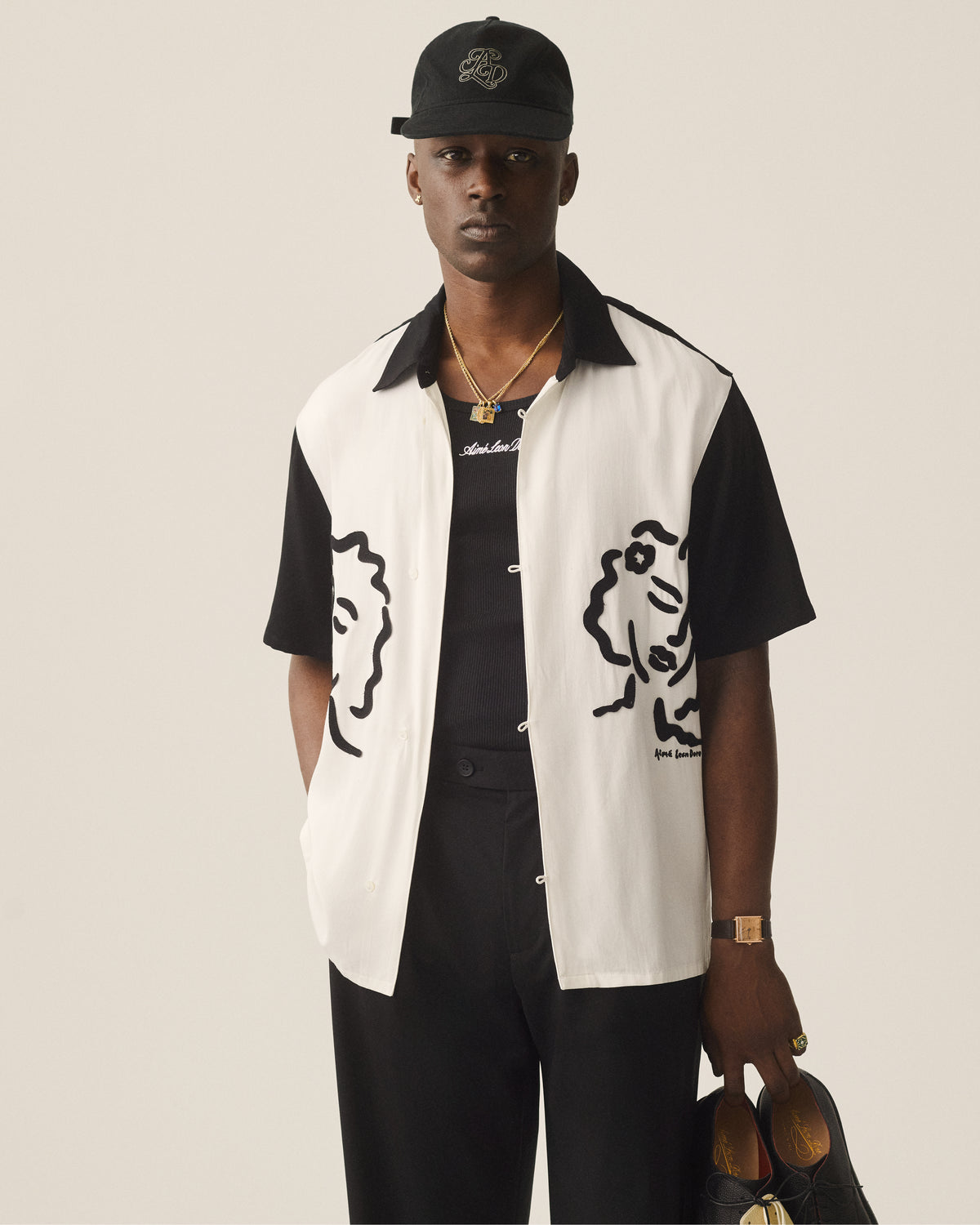 A man in black pants and a black cap wears an Embroidered Logo Tank Top and holds shoes.