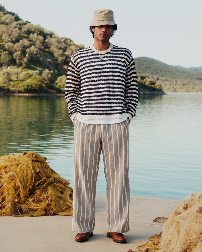Man in Wide Leg Shore Pant stands by water with fishing nets, trees, and hills in the background.