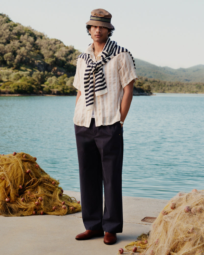 Man in a Linen Blend Striped Camp Shirt stands by a lake, dark pants on, fishing nets nearby.