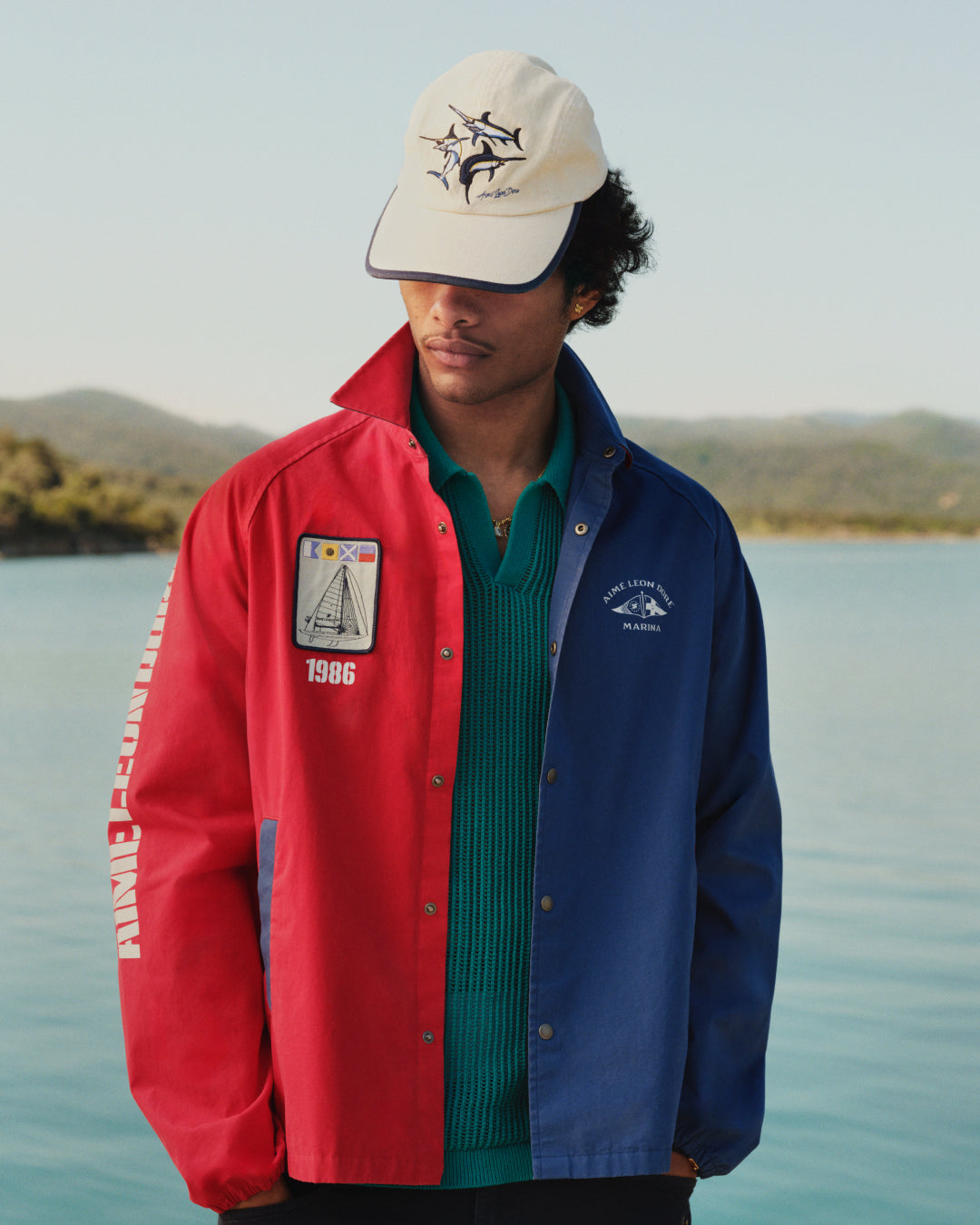 A man in a Souvenir Fishing Hat stands outdoors by water, looking down.