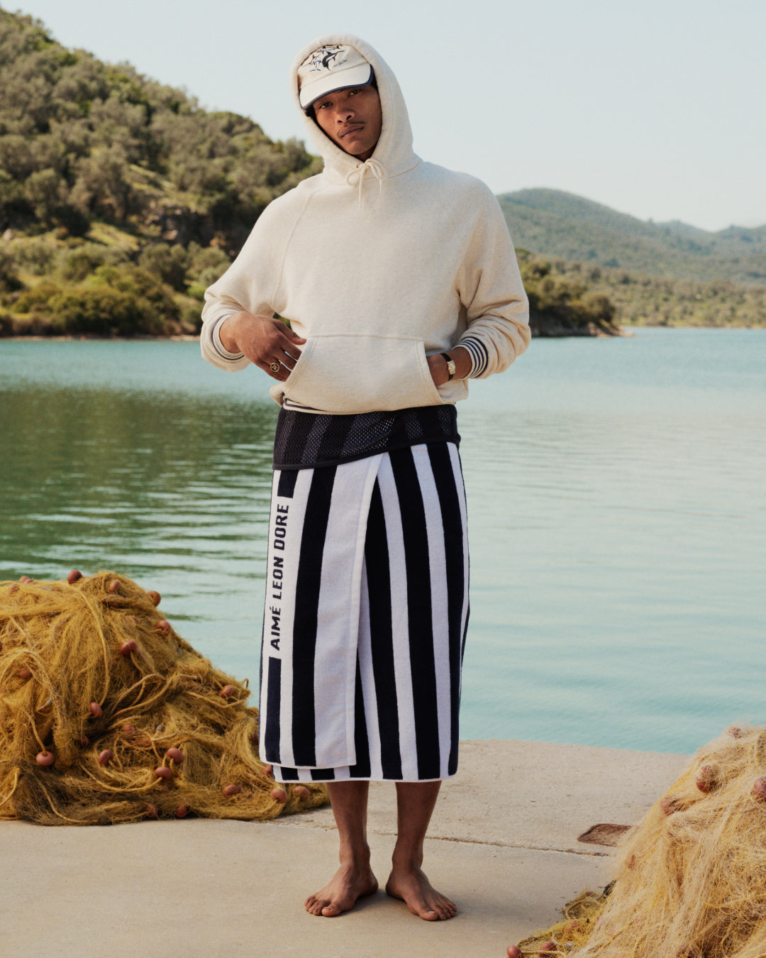 A man in a Dock Hoodie and cap stands barefoot on a dock by the lake, wearing a striped towel skirt.