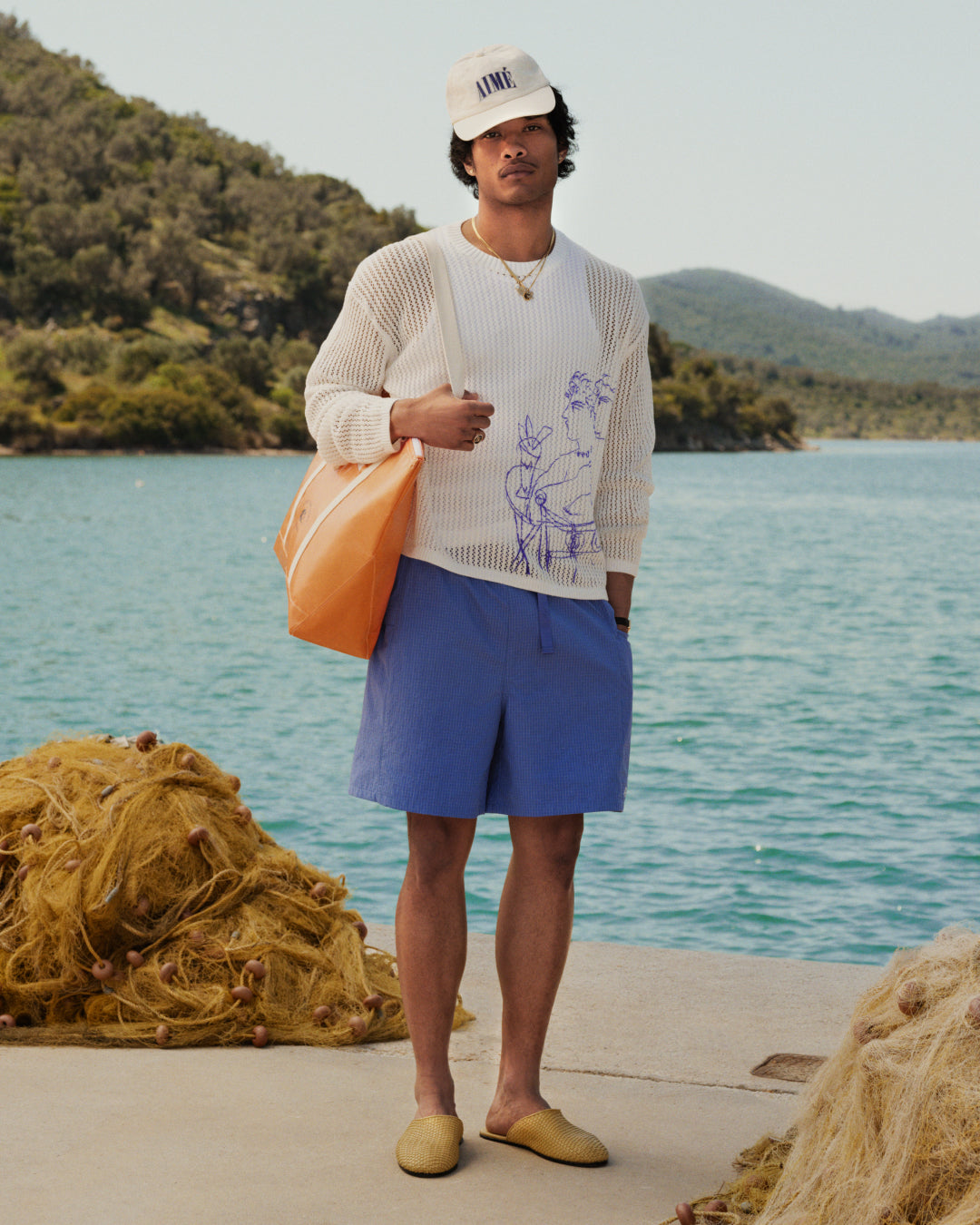 A man stands by water with a Carryall Marina Tote; nets, hills, and a duffel bag are in the background.