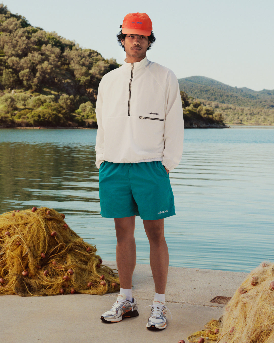 A person in a white jacket, orange cap, and Logo Swim Short stands by a lake with nets and trees in the background.