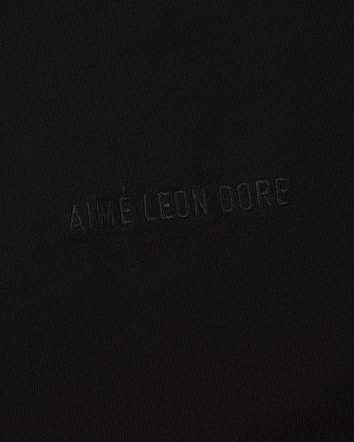 Close-up of Tonal Logo Tee's black fabric with AIMÉ LEON DORE subtly embossed in matching black lettering.