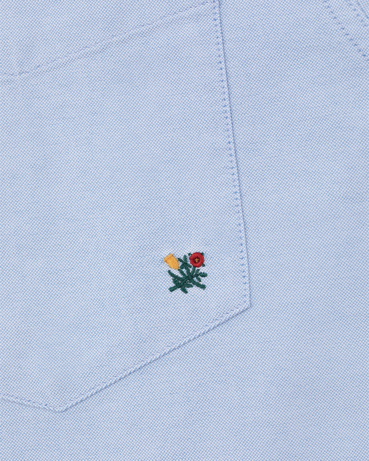 Light blue Crest Oxford Shirt with a fabric pocket featuring a small red and yellow flower embroidered near the bottom.