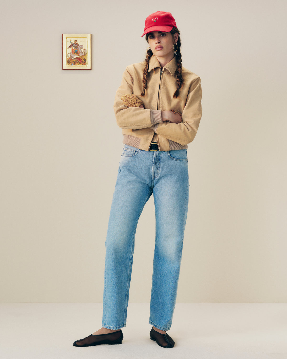 Woman in a red cap, tan jacket, blue jeans, and Mesh Ballet Flats stands indoors; art hangs on the wall behind her.